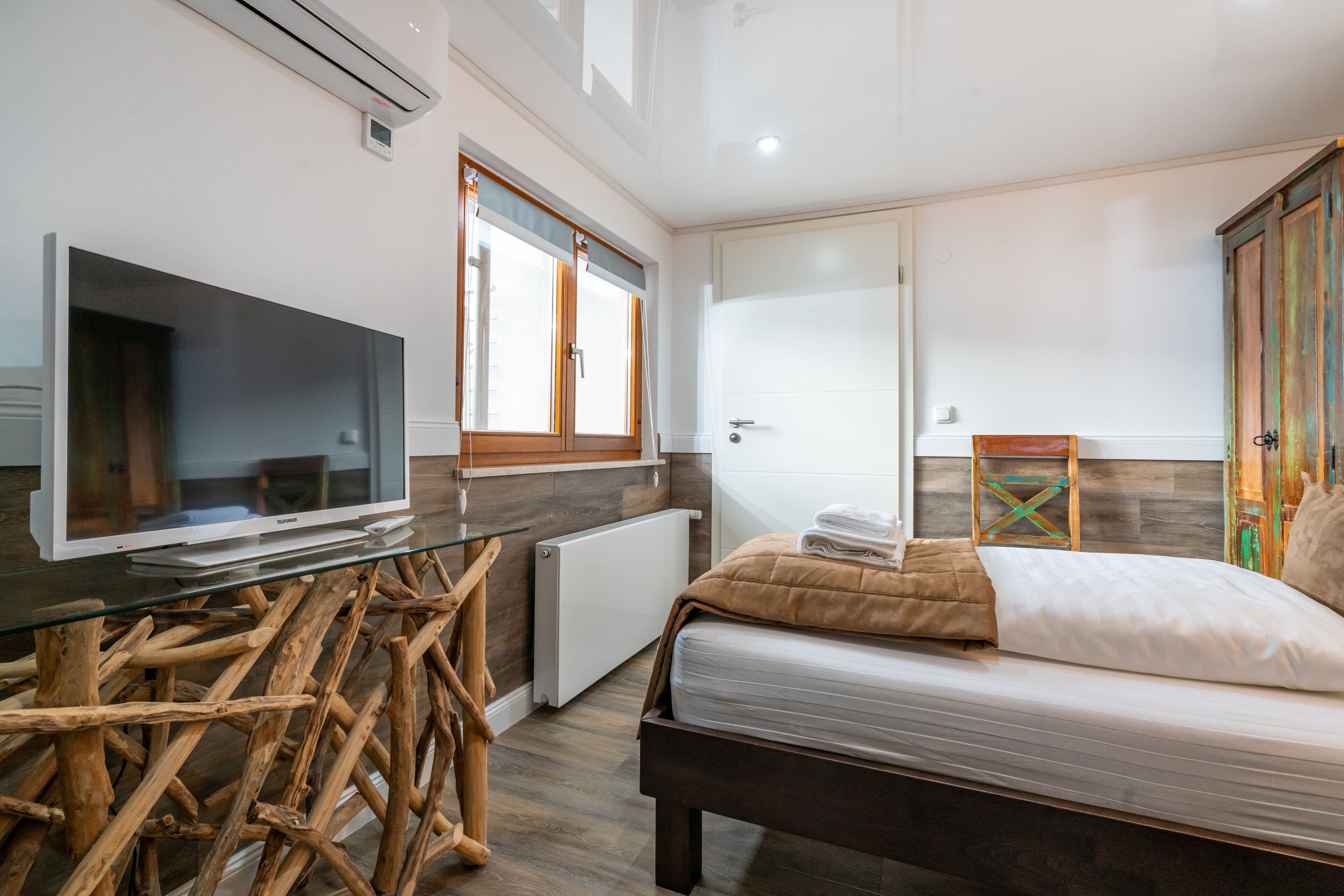 Un dormitorio moderno con una mesa de vidrio, muebles de madera, una cama con manta color beige y un televisor sobre un mueble bajo.