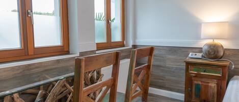 Una habitación con una mesa de comedor de madera, dos sillas, un mueble pequeño con una lámpara y ventanas amplias con marcos de madera.