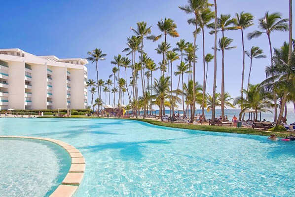 A resort pool with palm trees and a building in the background. - PanoramicHighView|BeachFront|FastWifi|FullKitchen (Juan Dolio)