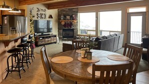 A cozy living room with a fireplace, a dining area with a round table, and a kitchen with a refrigerator and bar stools. - Schweitzer Condo. TRANSFERABLE SKI LIFT PASS INCLUDED! Hot Tub, 2-car Garage. (Sandpoint)
