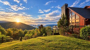 A house with a deck, surrounded by a fenced yard and trees, with a mountain backdrop and a clear sky.