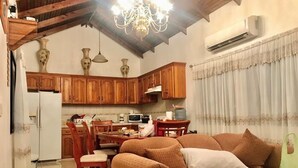 Una sala de estar acogedora con un sofá marrón, una mesa de comedor de madera y sillas. Se ve un área de cocina con armarios de madera y un refrigerador al fondo.