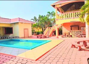 A pool area with a clear blue pool, a covered seating area with tables and chairs, and a building with a red-tiled roof. - Shores Plantation (Tela)
