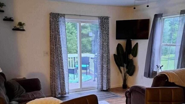 A living room with a dark wooden coffee table, a brown sofa, and a brown armchair. There is a ceiling fan, a flat-screen TV mounted on the wall, and a sliding glass door leading to an outdoor area with a table and chairs.