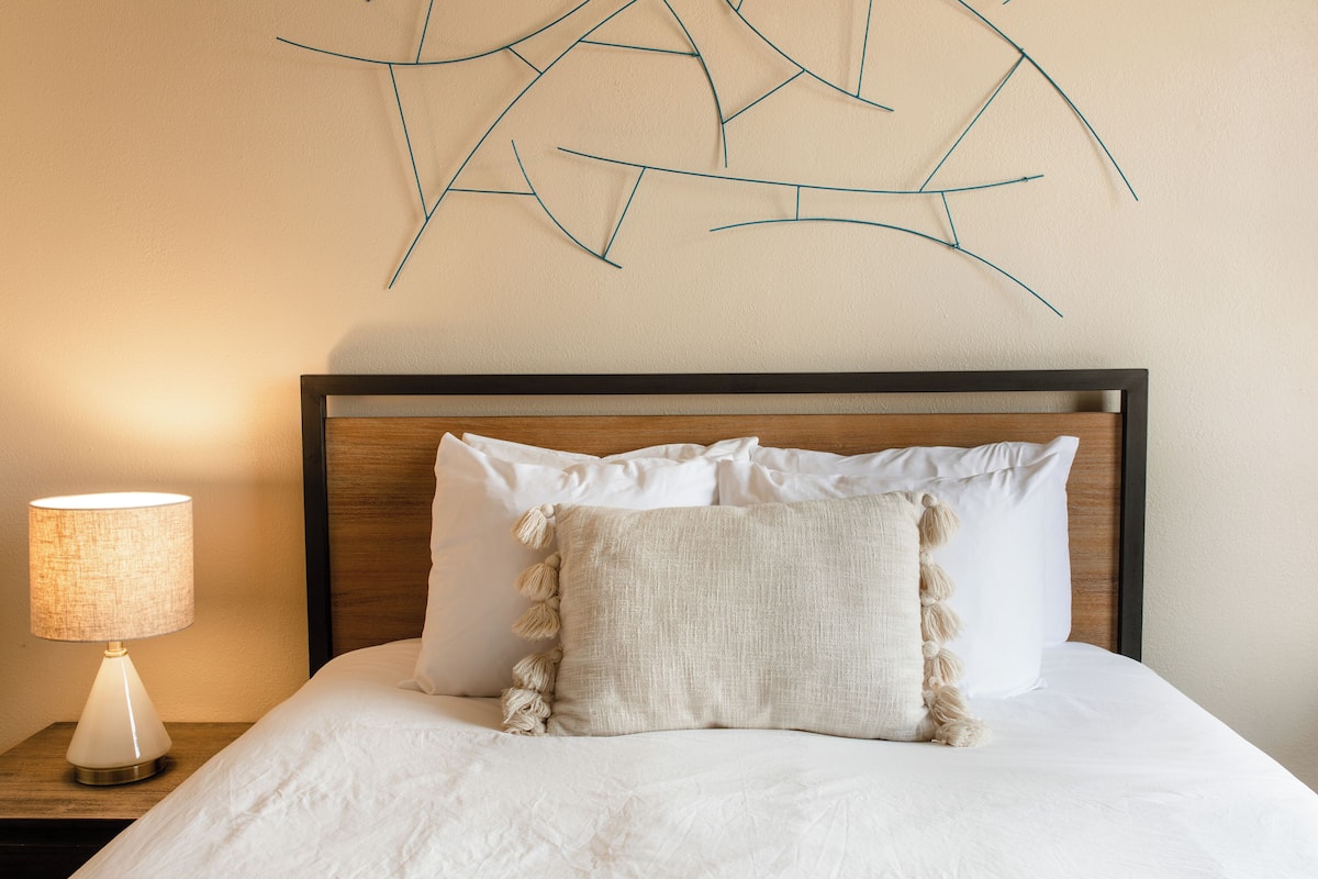 Cama bem arrumada com lençóis brancos e almofada decorativa texturizada, luminária de cabeceira em um criado-mudo de madeira e uma obra de arte na parede acima da cama.