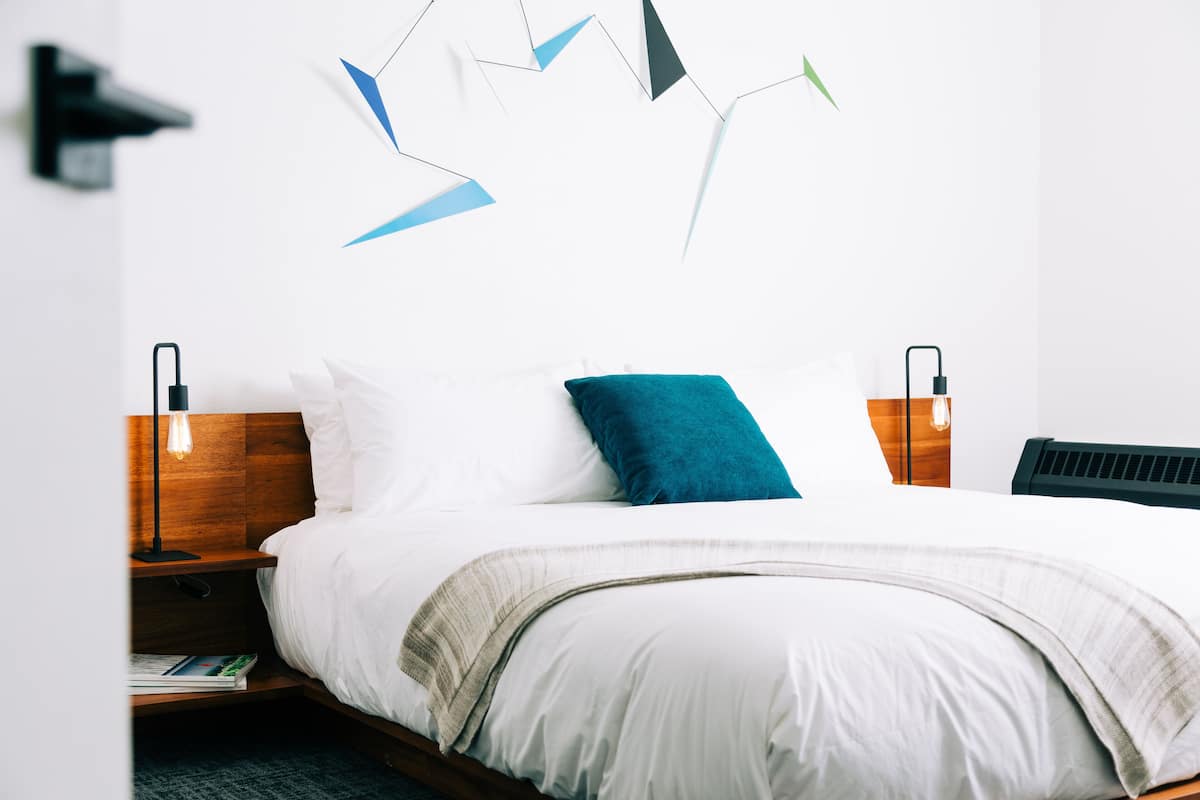 Quarto de hotel moderno com uma cama grande, cabeceira de madeira, duas mesas de cabeceira com abajures e uma obra de arte abstrata na parede.
