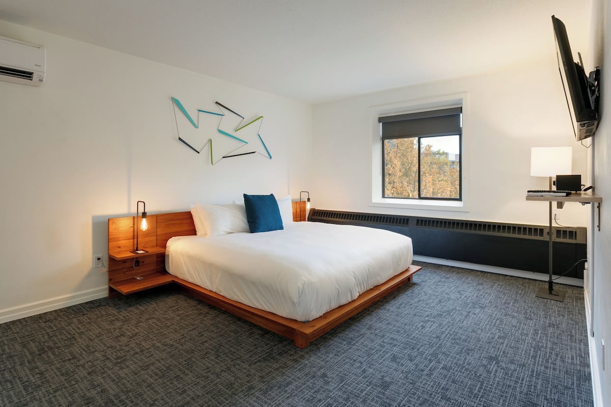 Quarto de hotel moderno com uma cama grande, uma mesa com computador, uma janela com vista e uma TV fixada na parede.