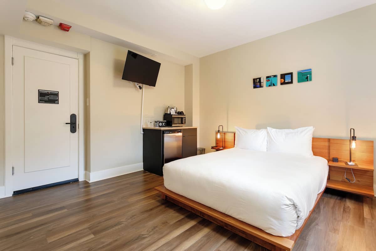 Quarto de hotel com uma cama grande, TV fixada na parede, pequeno espaço de cozinha e piso de madeira.