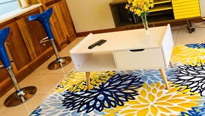 A modern living room with a white desk, two blue bar stools, a TV on the wall, a yellow and blue patterned rug, and a small vase with yellow flowers. - Waterloo Executive Guest Apartment (Kingston)