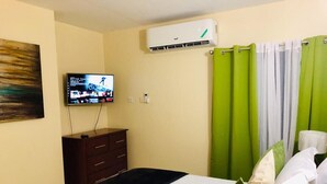 A hotel room with a bed, a TV mounted on the wall, a dresser, and a window with green curtains. - Waterloo Executive Guest Apartment (Kingston)