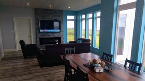 A modern living room with a fireplace, a large TV mounted on a stone wall, and a dining area with a wooden table and chairs. - Ocean Front Cottage. (Cable Head East)