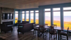 A spacious room with a large wooden ceiling, a fireplace, a television mounted on a brick wall, and a dining area with a view of the ocean. - Ocean Front Cottage. (Cable Head East)