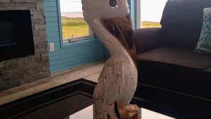 A room with a stone fireplace, a large window with a view of the outdoors, a dark-colored sofa, and a decorative wooden sculpture on a glass table. - Ocean Front Cottage. (Cable Head East)