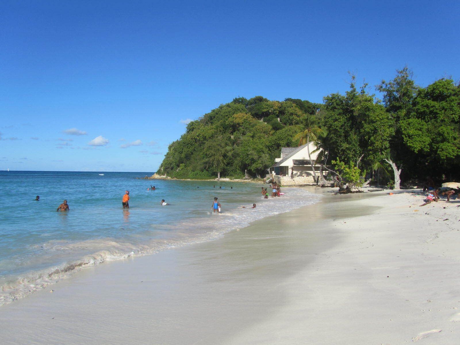 A beach with clear waters, people swimming and sunbathing, and a house on the shore.