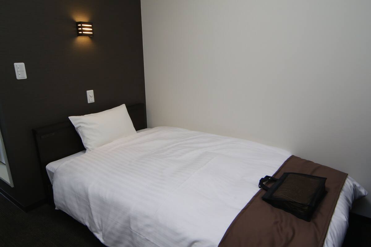 A neatly made bed with a white comforter and a brown blanket, a headboard, and a wall-mounted light fixture.
