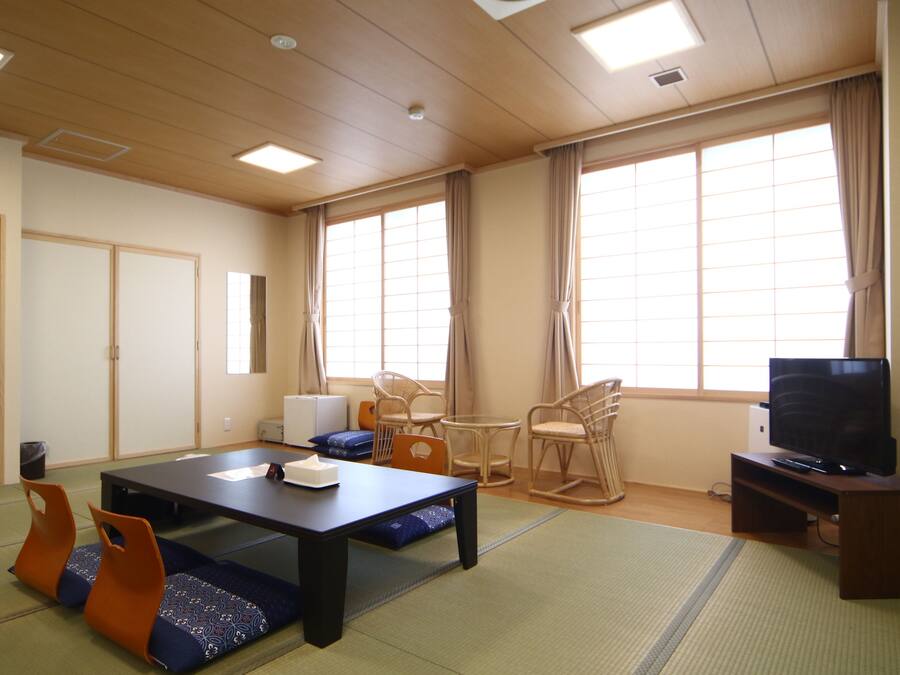 A room with tatami mats, a low table, chairs, a television, and large windows.