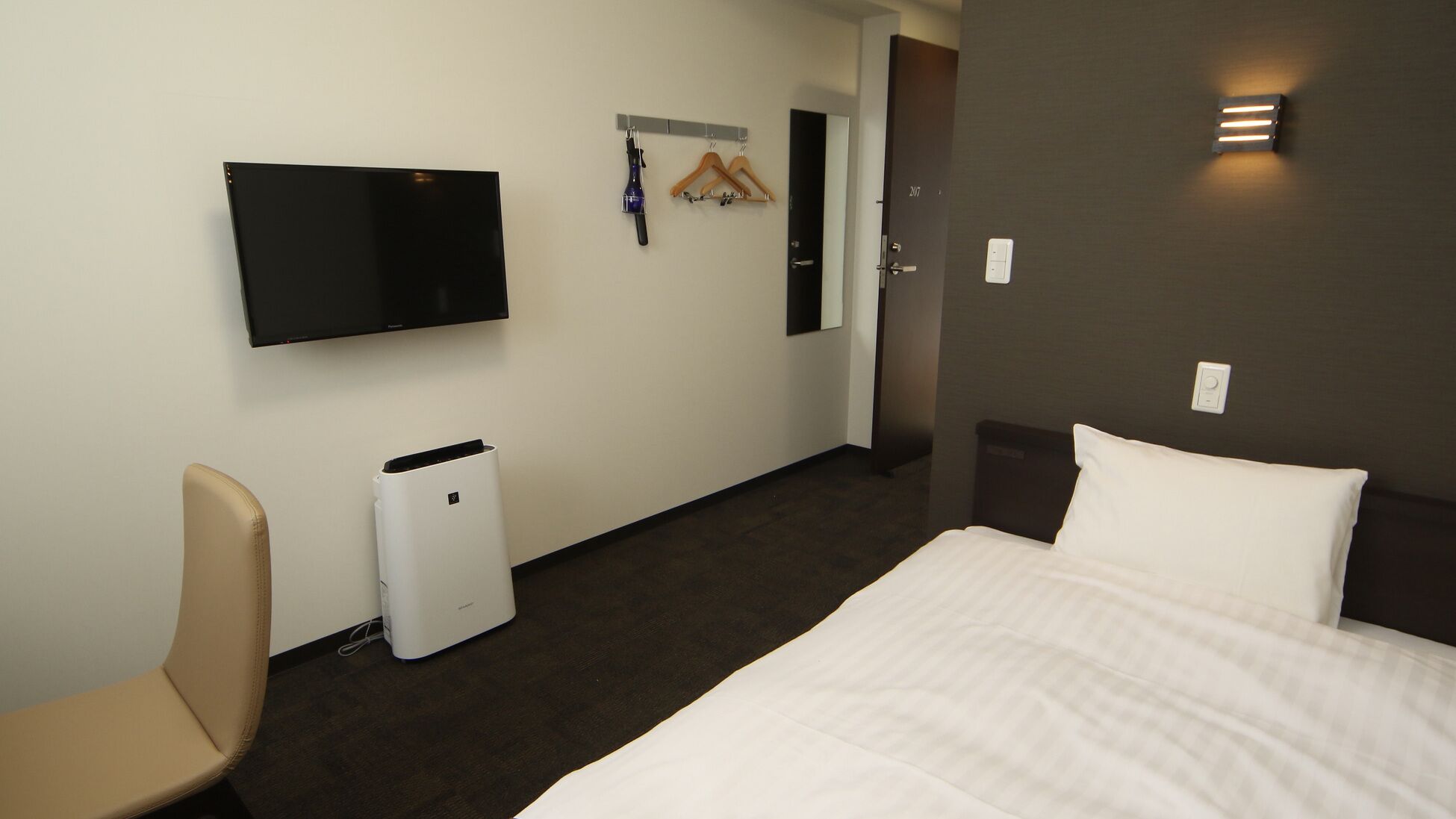 A hotel room with a bed, a chair, a TV, a coat rack, and a trash bin.