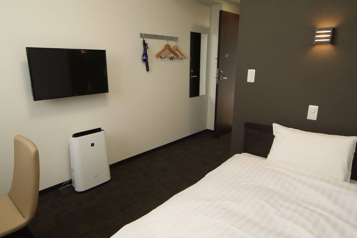 A hotel room with a bed, a chair, a TV, a coat rack, and a trash bin.