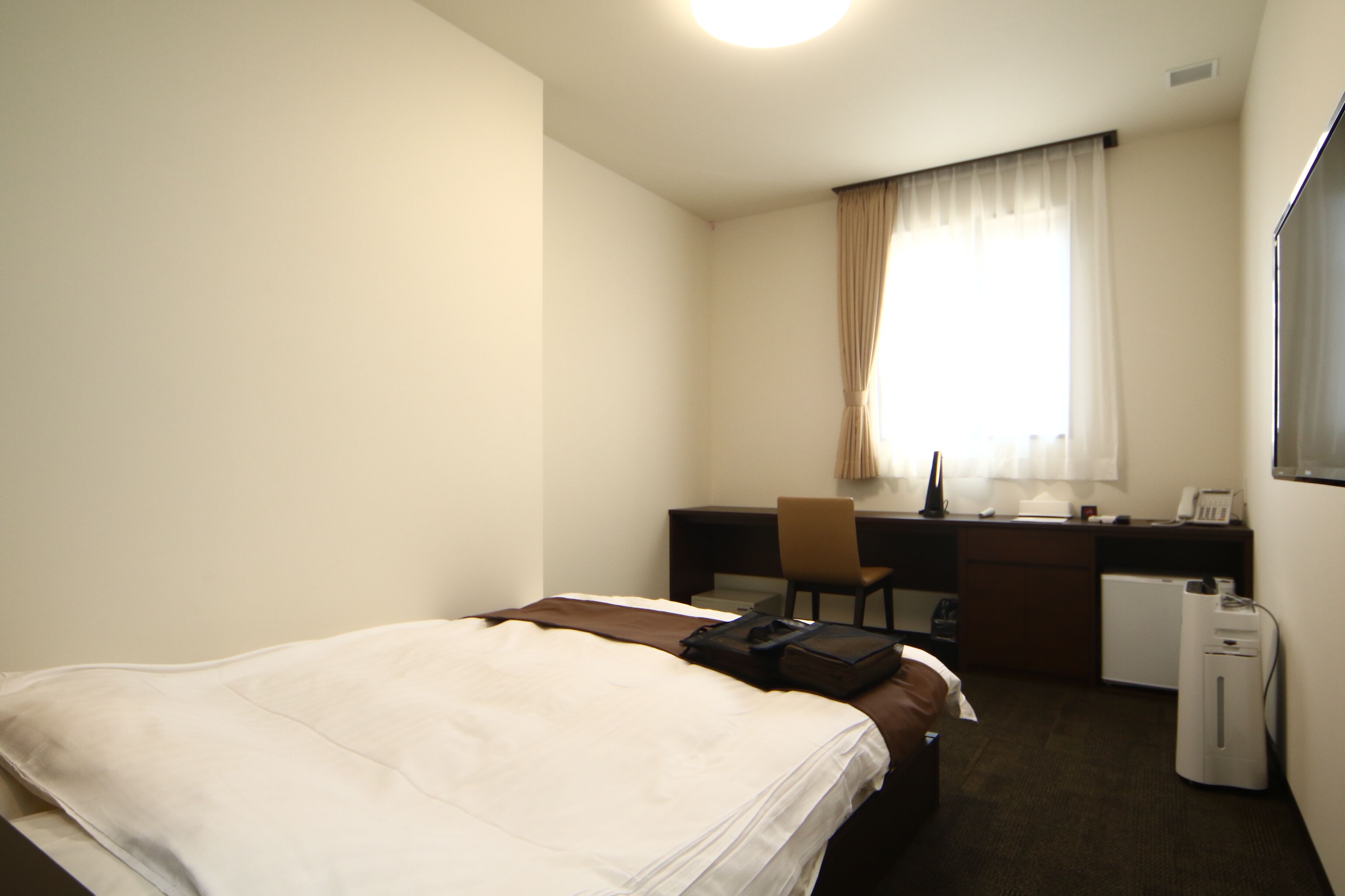 Una habitación de hotel con cama, escritorio, silla, refrigerador pequeño y ventana con cortinas.