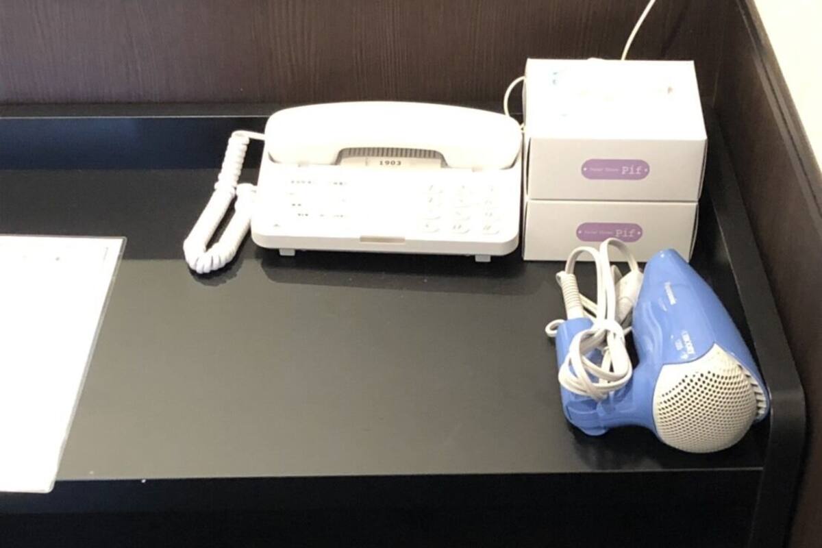 A desk with a white telephone, a blue hairdryer, a white mug, and a small trash bin.