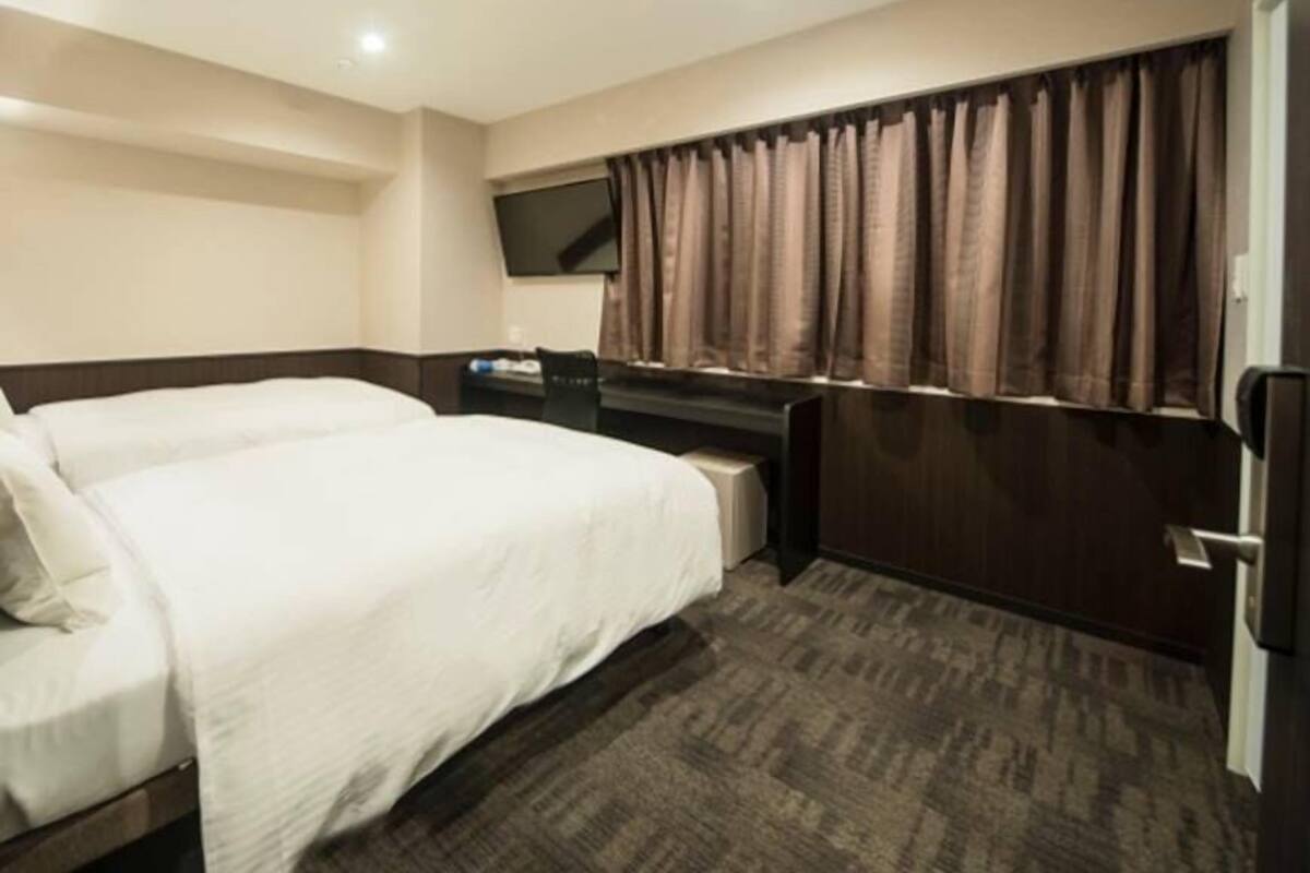 A hotel room with two beds, a desk, a television, and a window with curtains.