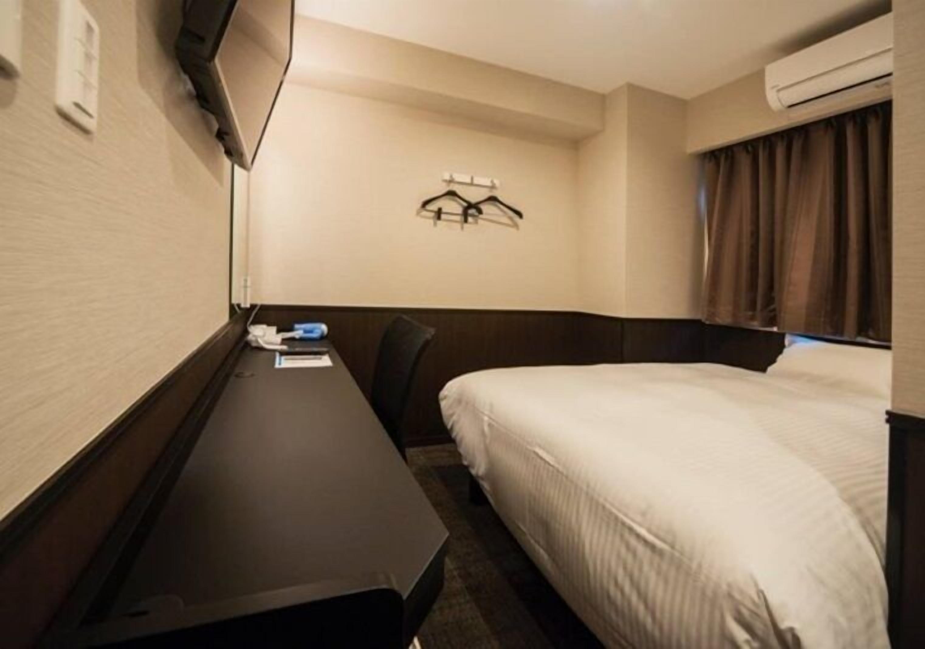 Quarto de hotel com cama, escrivaninha, cadeira, televisão fixada na parede e um cabideiro.
