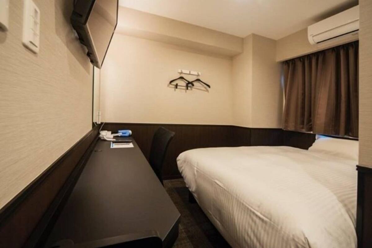 A hotel room with a bed, a desk, a chair, a TV mounted on the wall, and a coat rack.