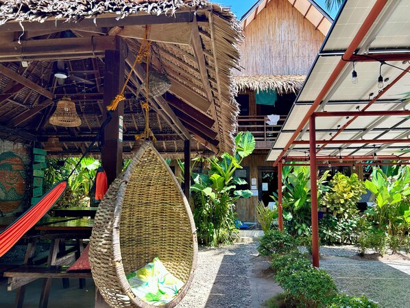 A rustic outdoor seating area with a hanging chair, wooden tables, and a thatched roof structure. - Ilakai Hostel - Adults Only (General Luna)