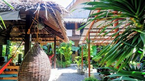 A traditional thatched-roof structure with a woven basket hanging from the ceiling, surrounded by lush tropical plants and a gravel pathway. - Ilakai Hostel - Adults Only (General Luna)
