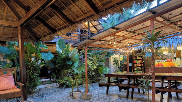 Un espacio de estar al aire libre con mesas y bancos de madera, rodeado de plantas en macetas y una estructura de techo de paja.