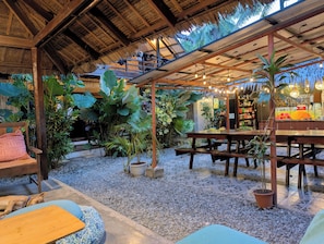An outdoor seating area with wooden tables and benches, surrounded by potted plants and a thatched roof structure. - Ilakai Hostel - Adults Only (General Luna)