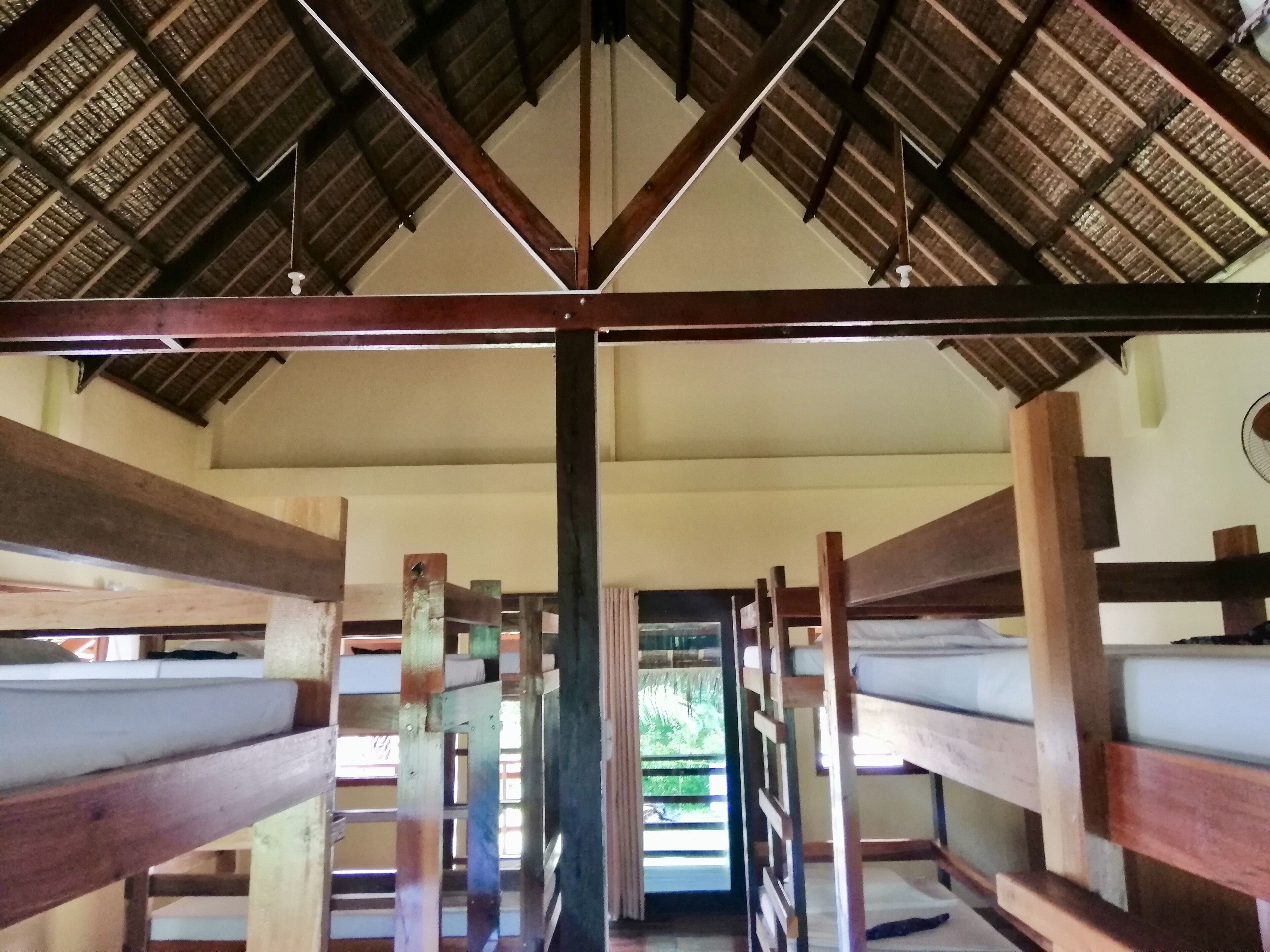 A room with bunk beds arranged in a row, wooden beams on the ceiling, and a view of greenery outside.