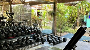 Gimnasio al aire libre con pesas y equipos de ejercicio variados, un área techada con techo de paja y un cartel que indica devolver las pesas después de usarlas.