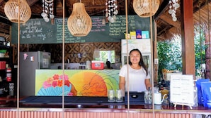 Una zona de bar con un menú en pizarra que enumera bebidas y precios, un refrigerador y un mural colorido en la pared.