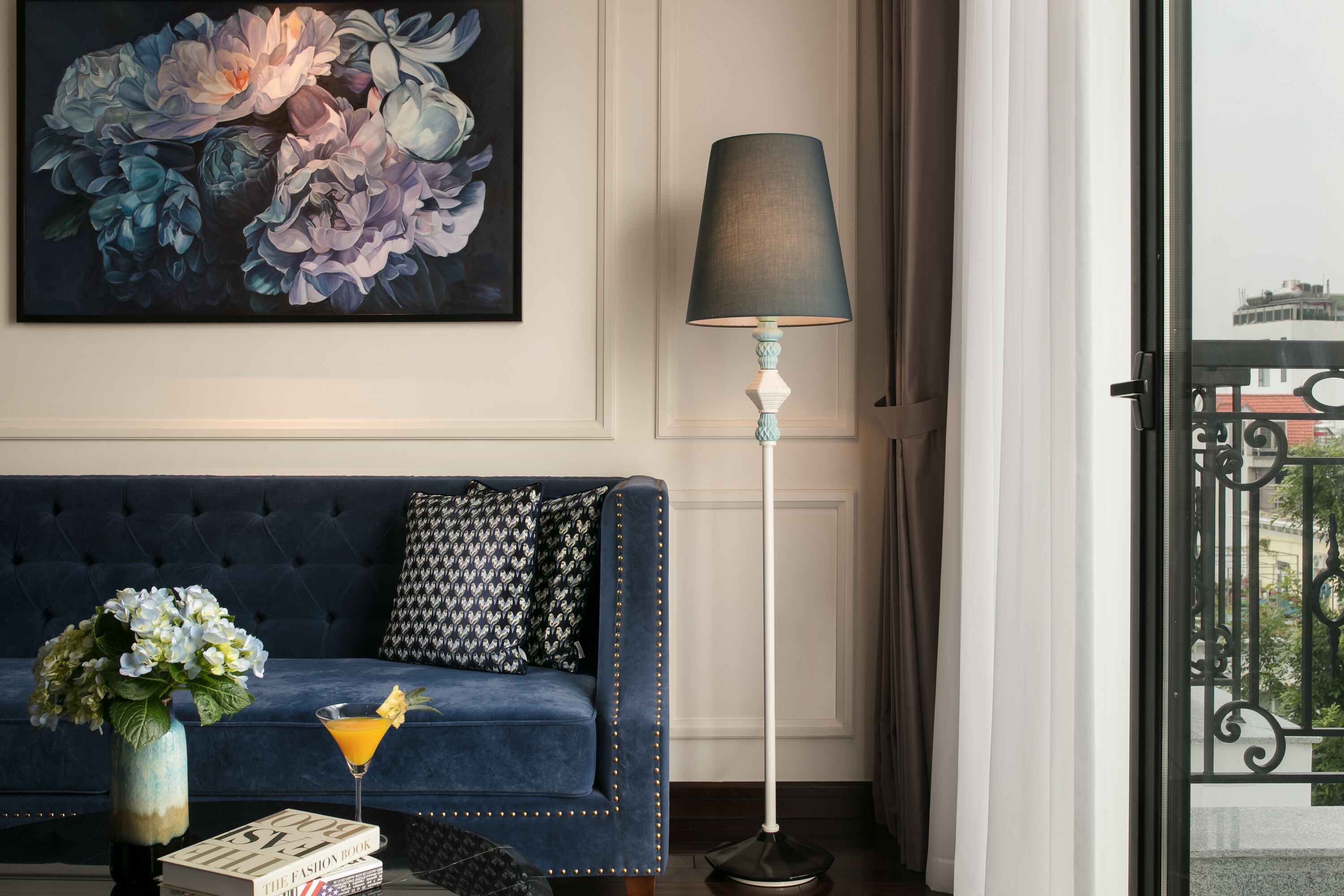 A dark blue sofa with patterned pillows, a floor lamp, a vase with flowers, a martini glass, and a book titled 'The Fashion Book' on a table.