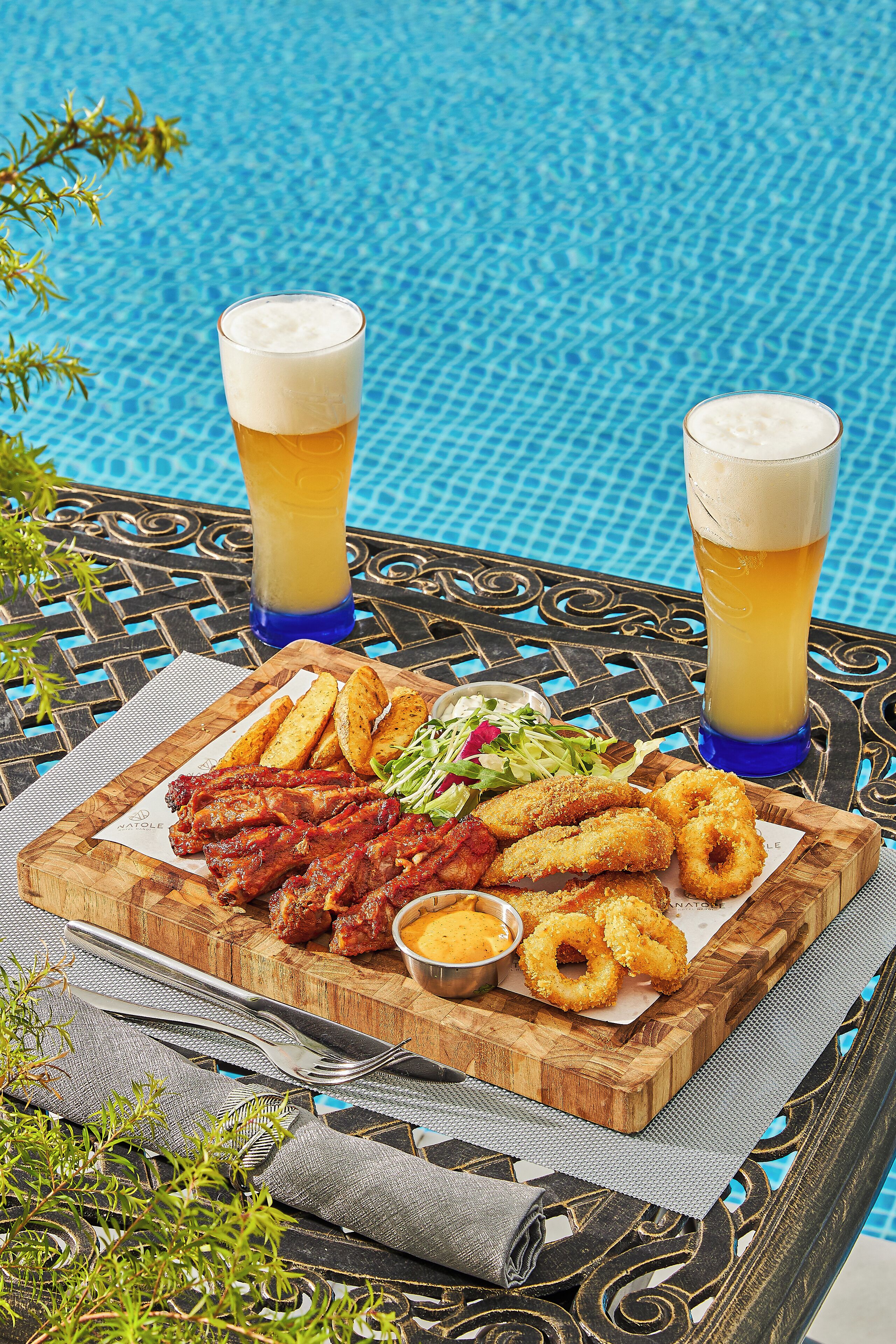 A dining table with a wooden board holding fried chicken, onion rings, and a salad, accompanied by two glasses of beer.