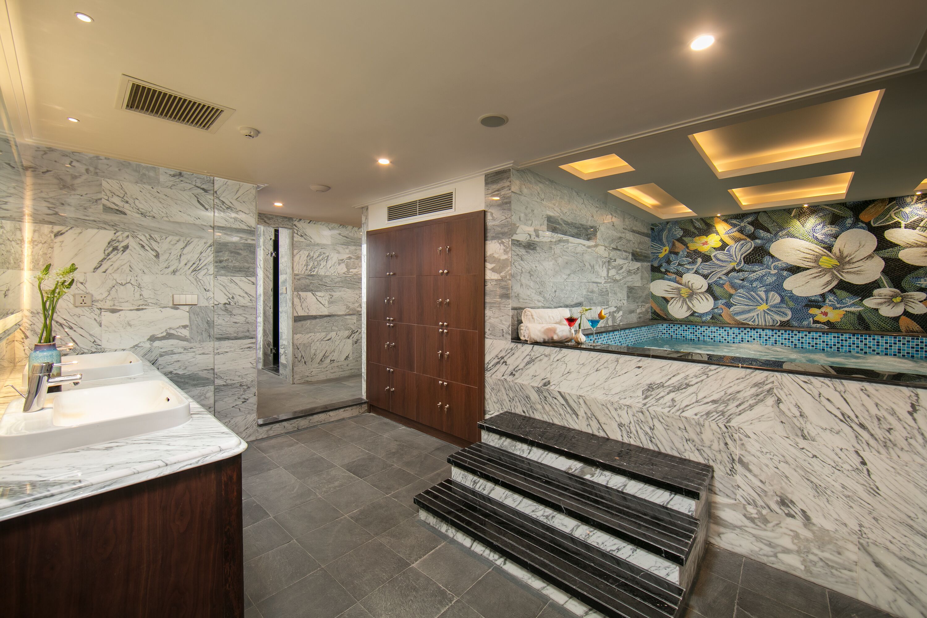 A modern bathroom with a marble sink, wooden cabinets, a shower area, and a decorative wall with floral tiles.