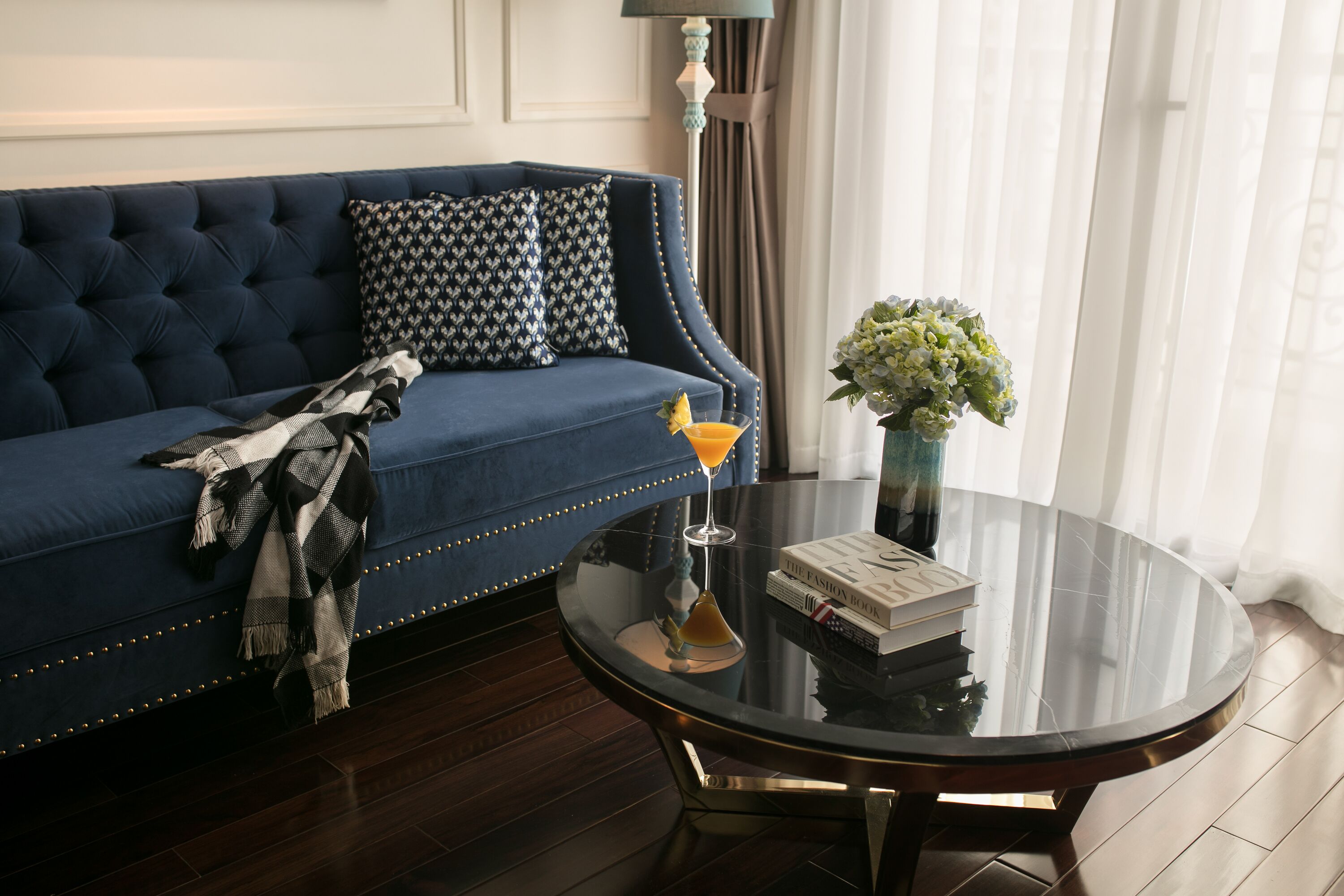 A blue sofa with patterned pillows, a glass table with a book and a vase of flowers, and a lamp in the background.
