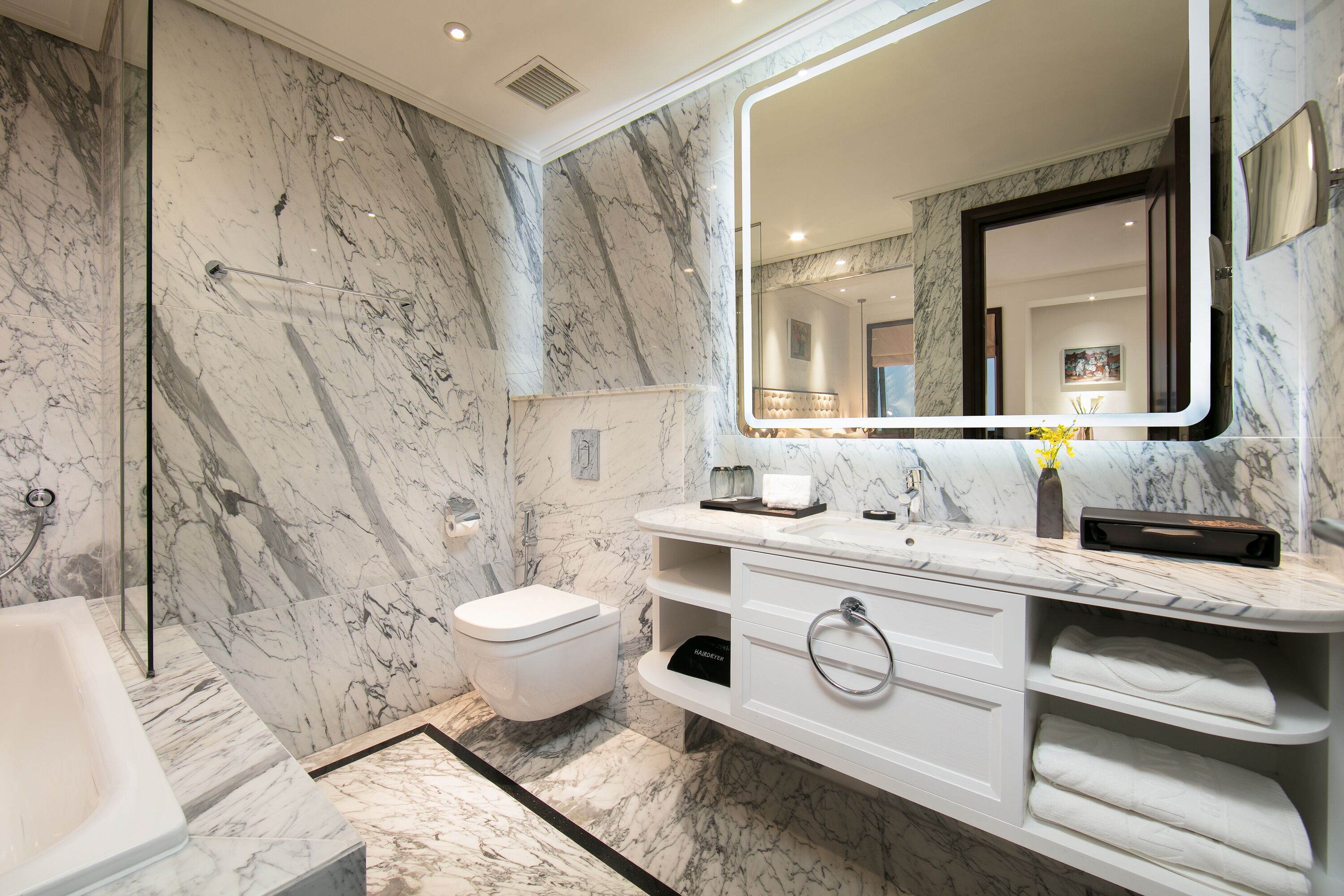 A bathroom with a marble wall, a glass shower enclosure, a white toilet, a bathtub, a vanity with a mirror, and a towel rack.