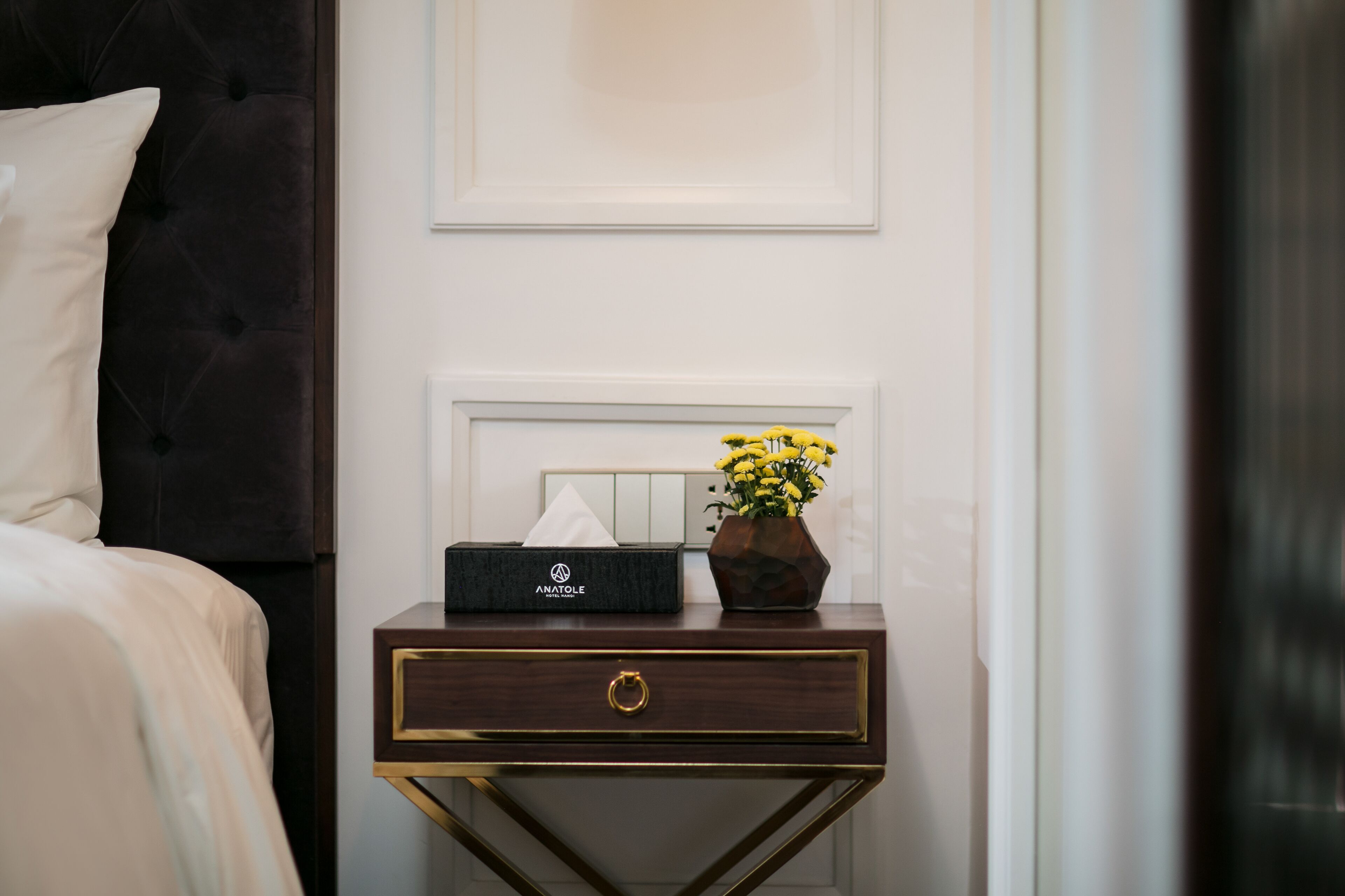 A neatly made hotel room with a bed, a bedside table with a tissue box and a vase of flowers, and a wall-mounted light switch.