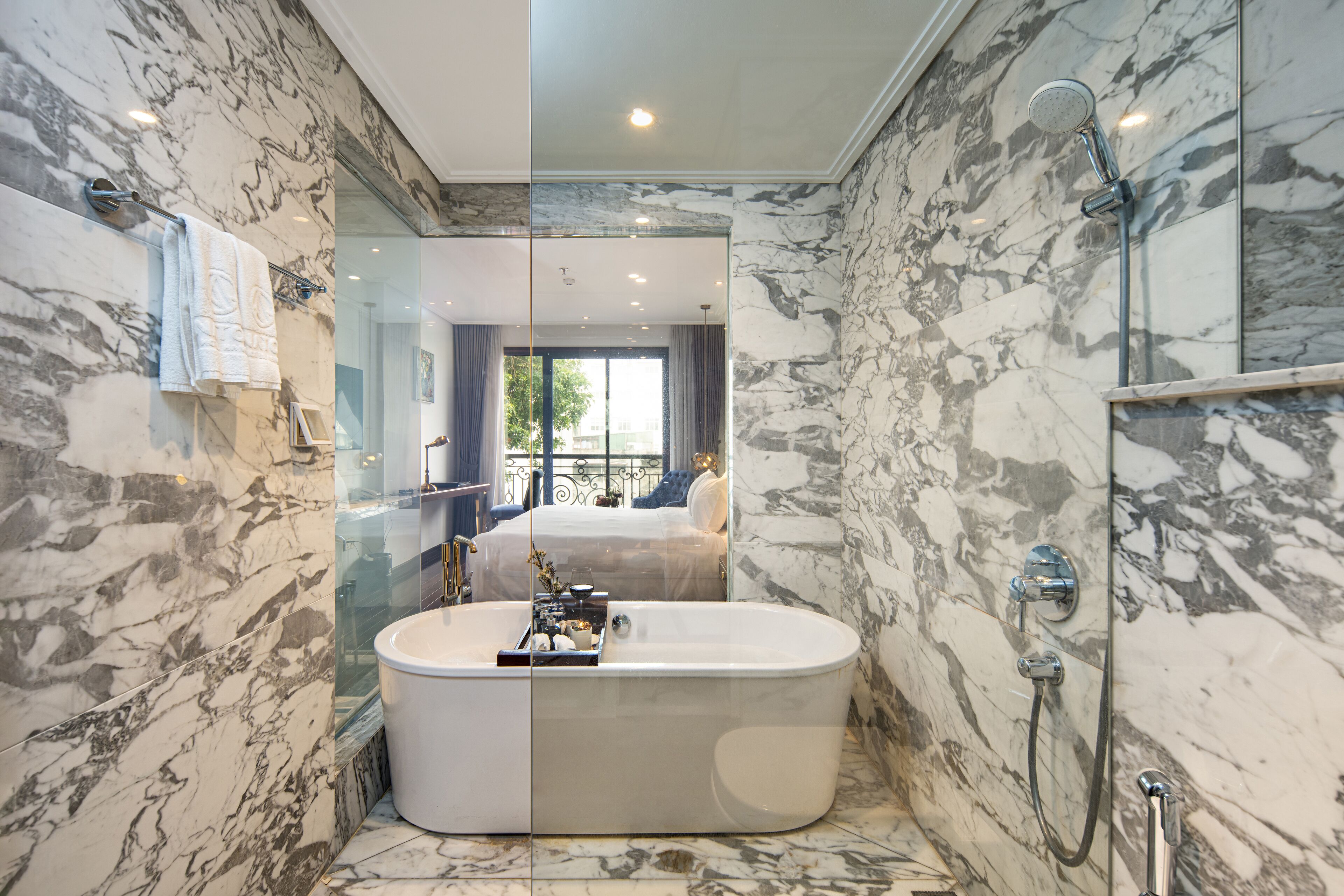 Un baño moderno con una bañera grande, zona de ducha con mampara de cristal, paredes de mármol y una habitación visible con cama y cortinas.