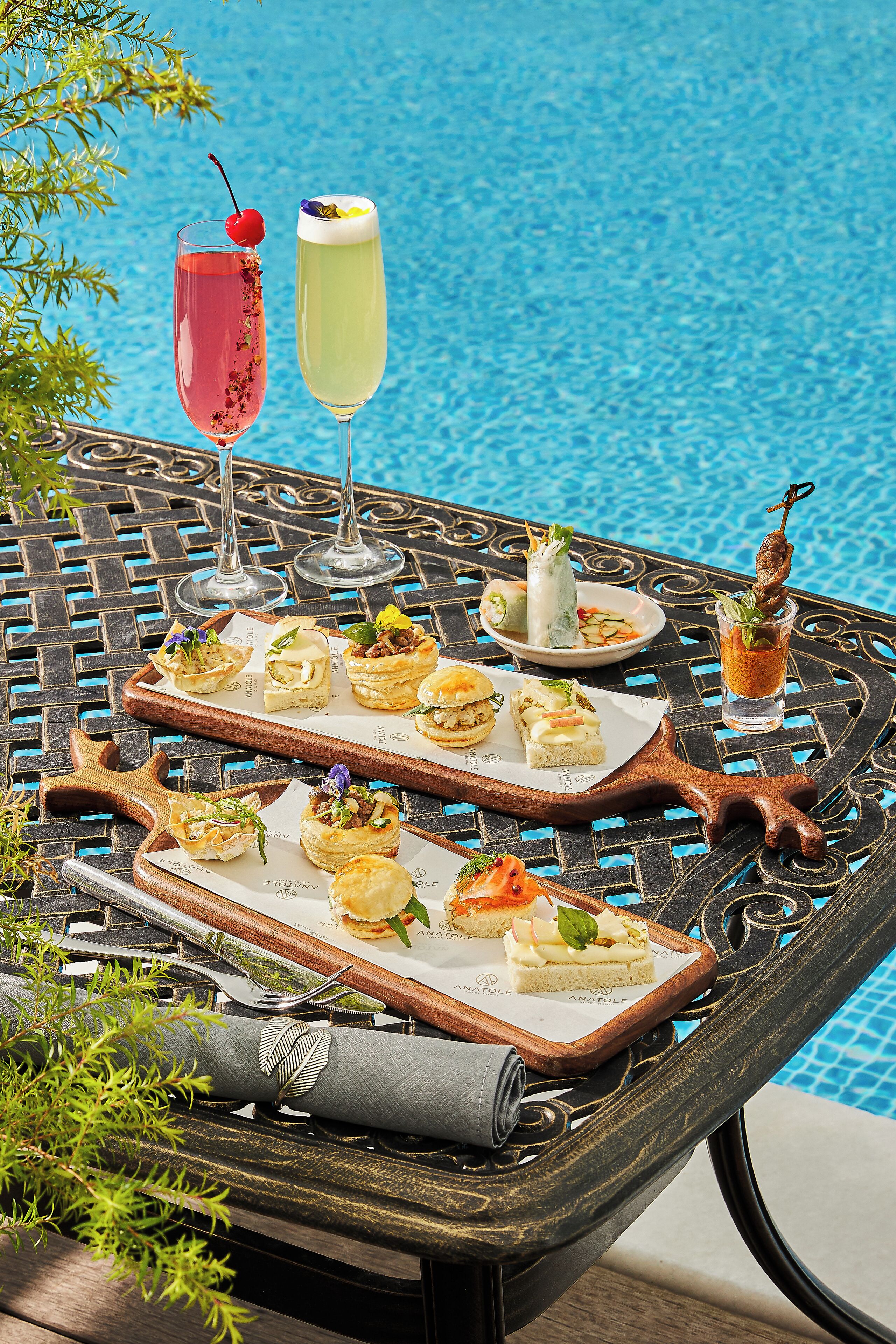 A table with various appetizers and drinks by a poolside, featuring a decorative tray and a rolled napkin.