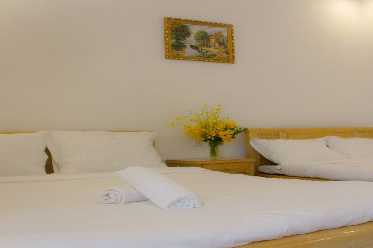 Quarto de hotel com duas camas, roupa de cama branca, cabeceira de madeira, mesa de cabeceira com um vaso de flores amarelas e uma moldura na parede.