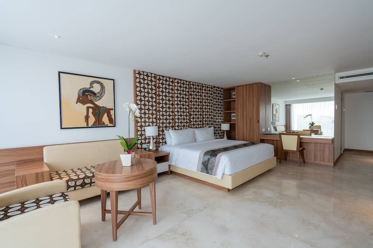 A modern hotel room with a bed, a desk, a chair, a small table with a plant, and a decorative wall panel.