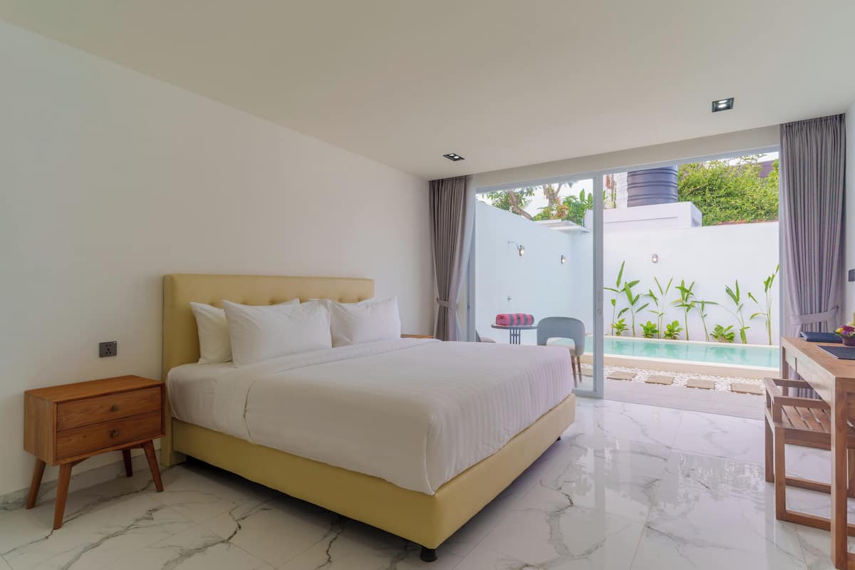 A bedroom with a bed, a nightstand, a table, and a view of a swimming pool and greenery outside.