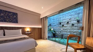 A hotel room with a bed, a nightstand with a lamp, a wooden chair, a small table, and a view of a stone wall and greenery outside. - The Capital Hotel and Resort Seminyak (Keroboken Kelod)
