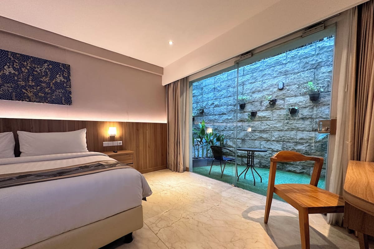 A hotel room with a bed, a nightstand with a lamp, a wooden chair, a small table, and a view of a stone wall and greenery outside.