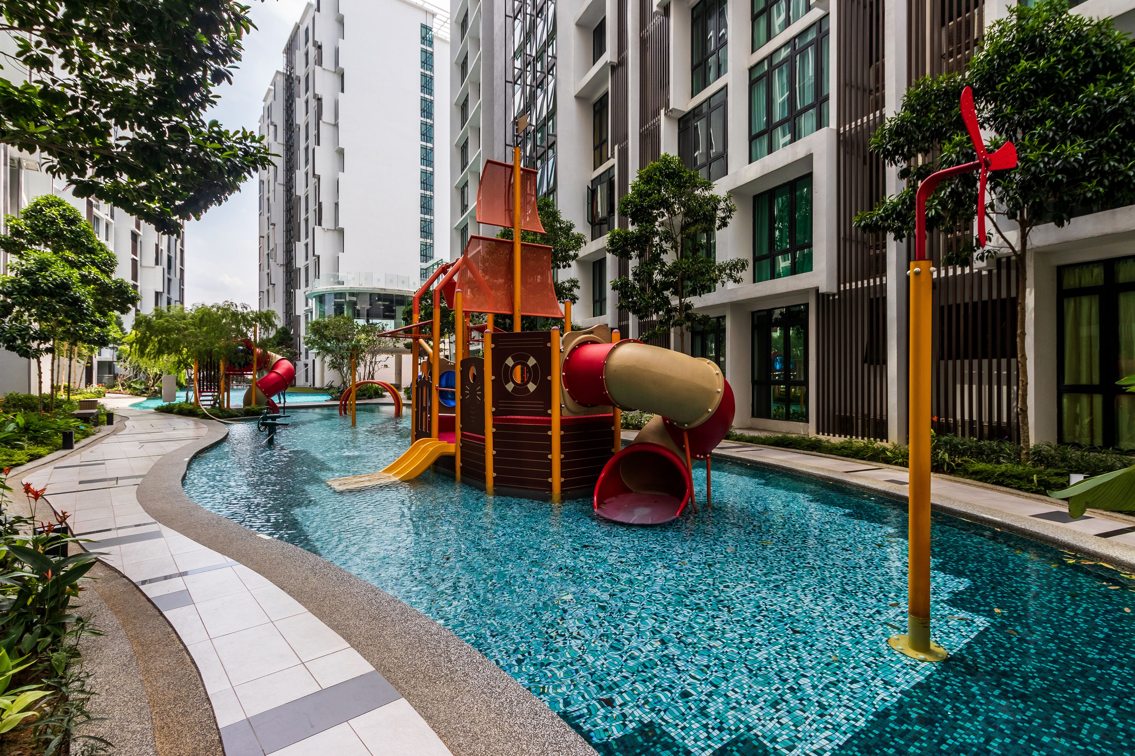 Un área de piscina con un parque infantil, rodeada de edificios modernos y vegetación.