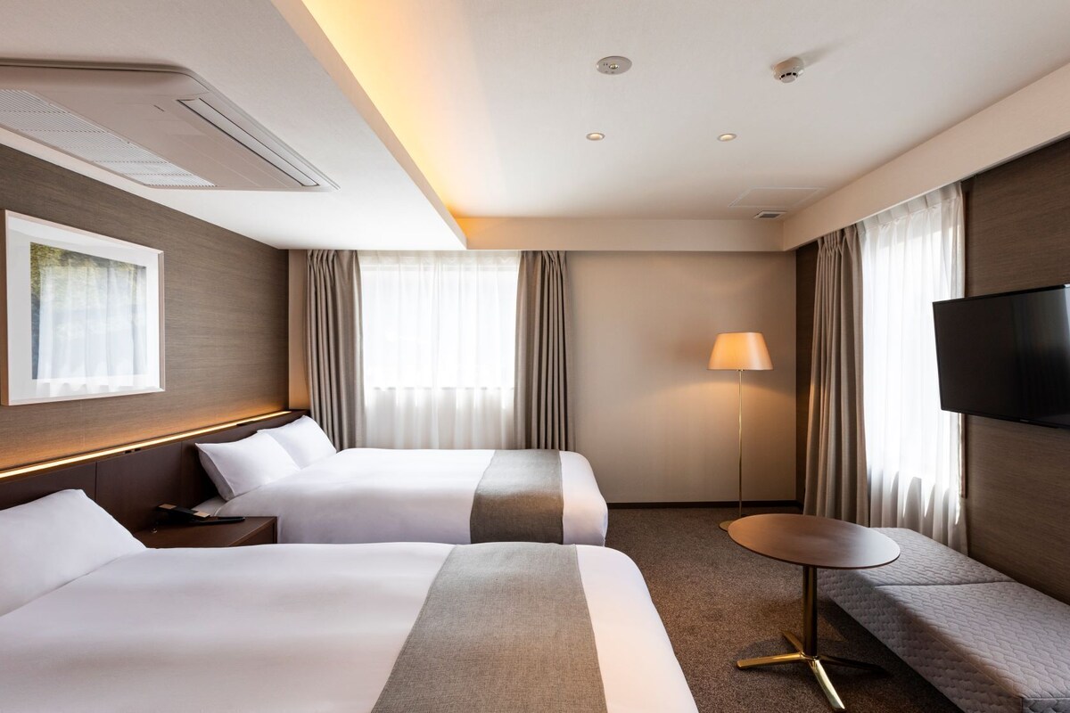 Habitación de hotel con dos camas, televisión, lámpara y ventana con cortinas.