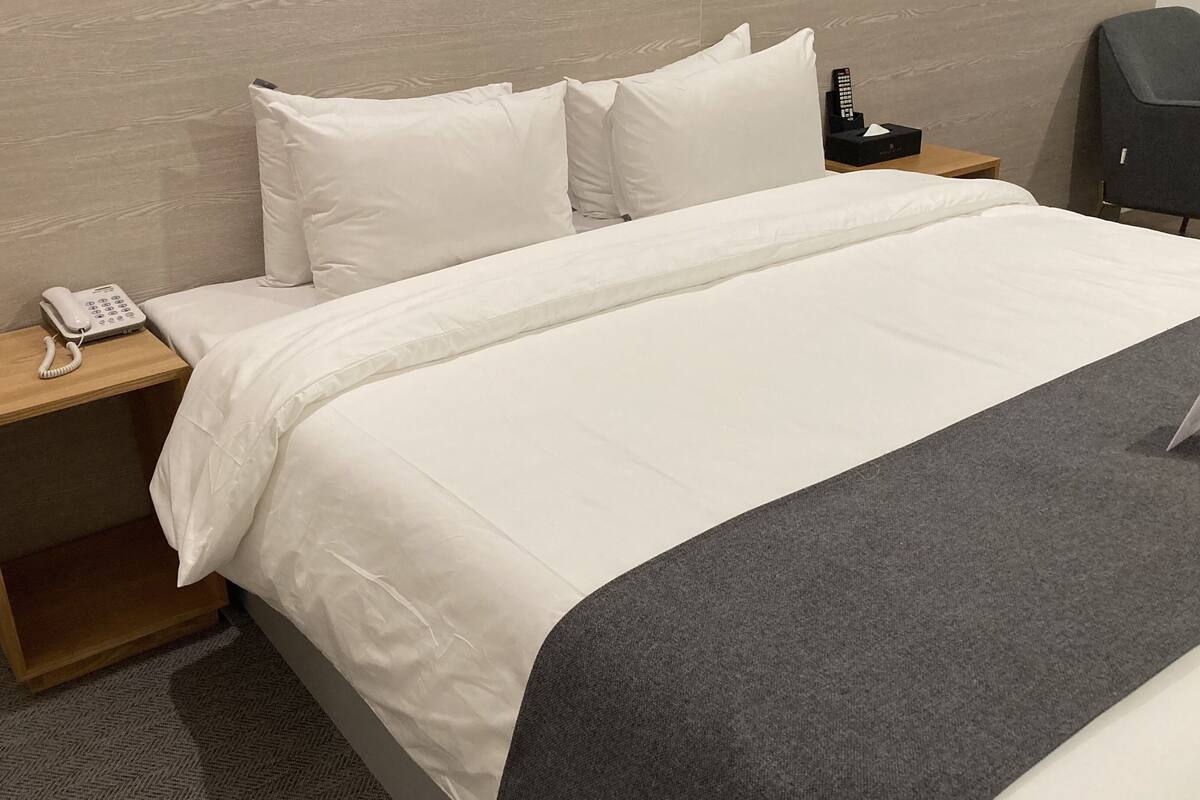 Una cama bien hecha con sábanas blancas, una mesita de noche de madera con un teléfono blanco y un control remoto negro sobre la mesita.